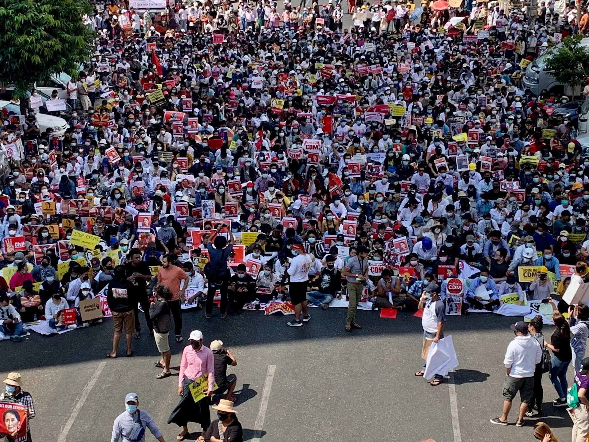 On February 17, 2021, at 11:53 AM, a large crowd of protesters was seen occupying the entire road on Anawrahta Road near Bogyoke Pagoda Road in Yangon. The crowd is sitting on the road, holding various signs and placards.Some signs include messages supporting the Civil Disobedience Movement (CDM) and calling for justice.Many participants are wearing face masks, hats, and helmets, indicating a concern for safety and protection.